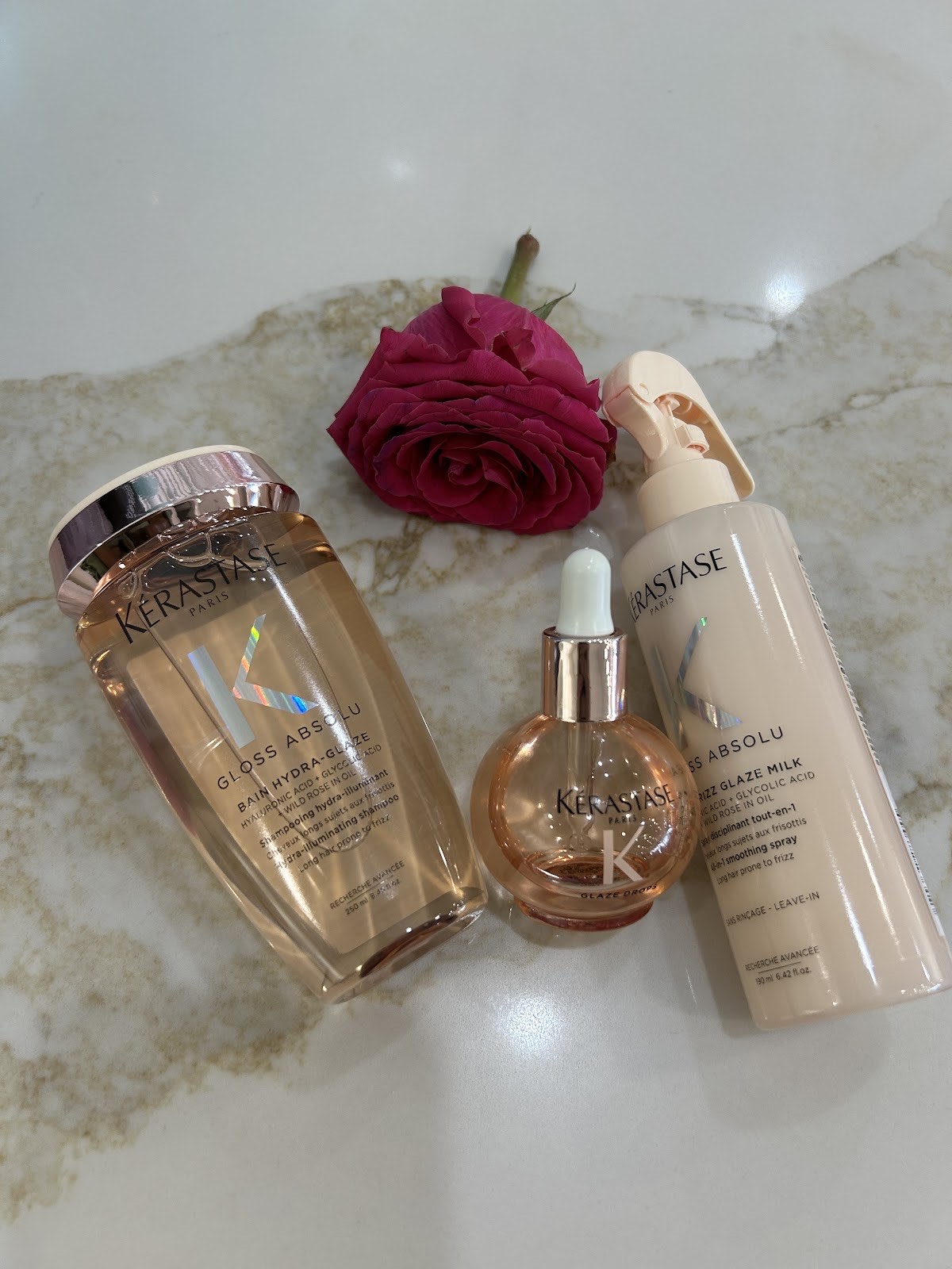 Kérastase Gloss Absolu shampoo, oil and milk on a marble surface beside a deep pink rose